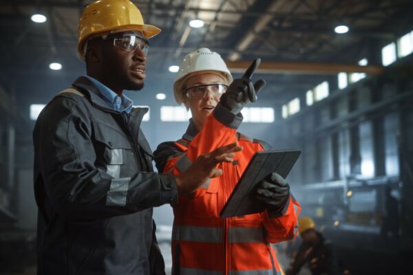 Two engineers with helmets and a tablet discuss work in an industrial setting.