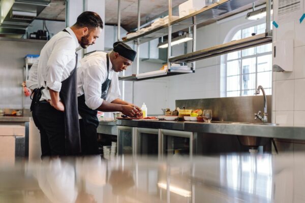 Two chefs in white shirts and black aprons work at a steel countertop in a bright, clean kitchen.
