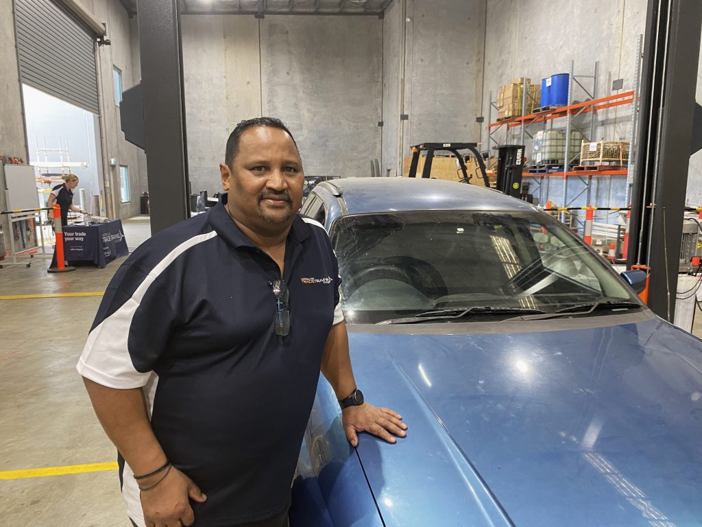 Man with car - no logo: Man standing next to a blue car in workshop, wearing a black and white polo shirt.