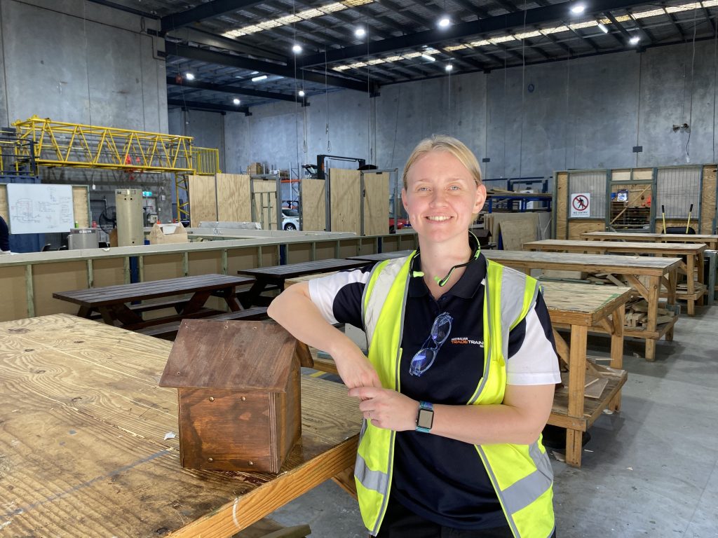 Smiling woman in safety vest posing with a wooden project in a large workshop.