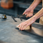 Hands using a caliper to measure a metal sheet on a workbench.