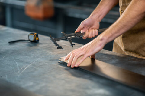 Hands using a caliper to measure a metal sheet on a workbench.