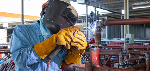 Welder in protective gear welding pipes with flying sparks.