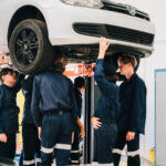 Mechanics in blue uniforms working under a car on a lift.