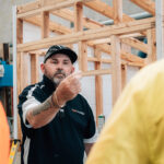 Instructor in a black polo gesturing toward a wooden house frame.