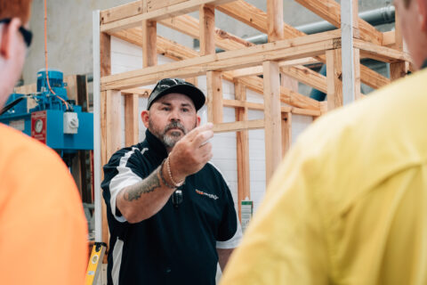 Instructor in a black polo gesturing toward a wooden house frame.