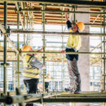 Two workers in high-vis gear assembling scaffolding on a site.