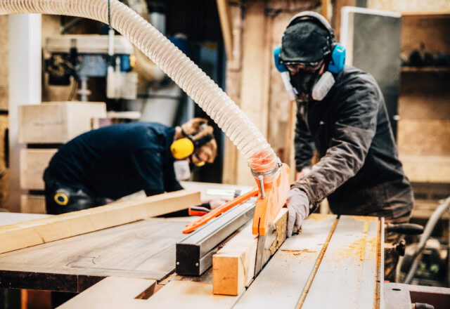 Woodworkers cutting lumber on a table saw with safety gear.