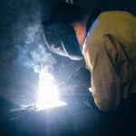 Welder in safety gear working on metal with bright blue sparks.