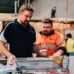 Two men in work gear inspecting an open electrical control box.