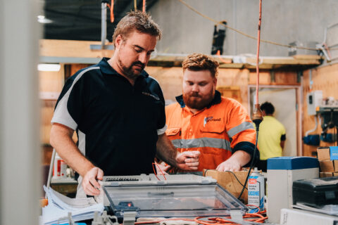 Two men in work gear inspecting an open electrical control box.