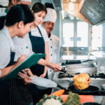 Culinary student frying food while others watch and take notes.