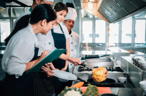 Culinary student frying food while others watch and take notes.